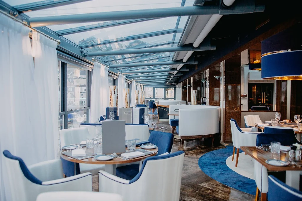 The image shows a luxurious restaurant with a modern design, featuring a glass ceiling, blue and white furniture, and a blue rug. The restaurant is well-lit, with large windows offering a view of the cityscape. The layout is organized, with tables and chairs arranged for guests. The restaurant is ready to serve, and the image captures the elegance and sophistication of the space.