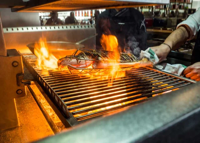 A chef in a black apron is grilling a large, pinkish salmon on a metal grill with flames. The grill is on a metal counter, and the background shows other grills and shelves with various items. The scene captures the essence of a professional kitchen, showcasing the chef's skill and dedication to the art of cooking.