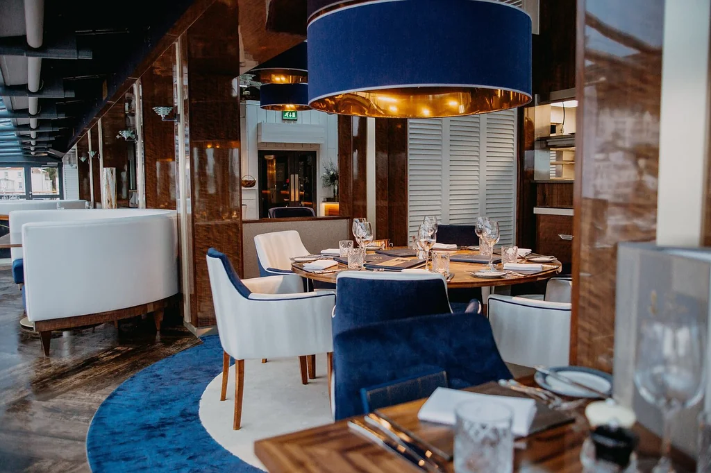 The image shows a luxurious restaurant with a modern, minimalist design. The main focus is a round table with a blue armchair, surrounded by white booths and a blue rug. The table is set with silverware and wine glasses, and the ceiling features a gold chandelier. The restaurant is well-lit, with a window on the right side allowing natural light. The arrangement of objects and the color scheme create a harmonious and inviting atmosphere.