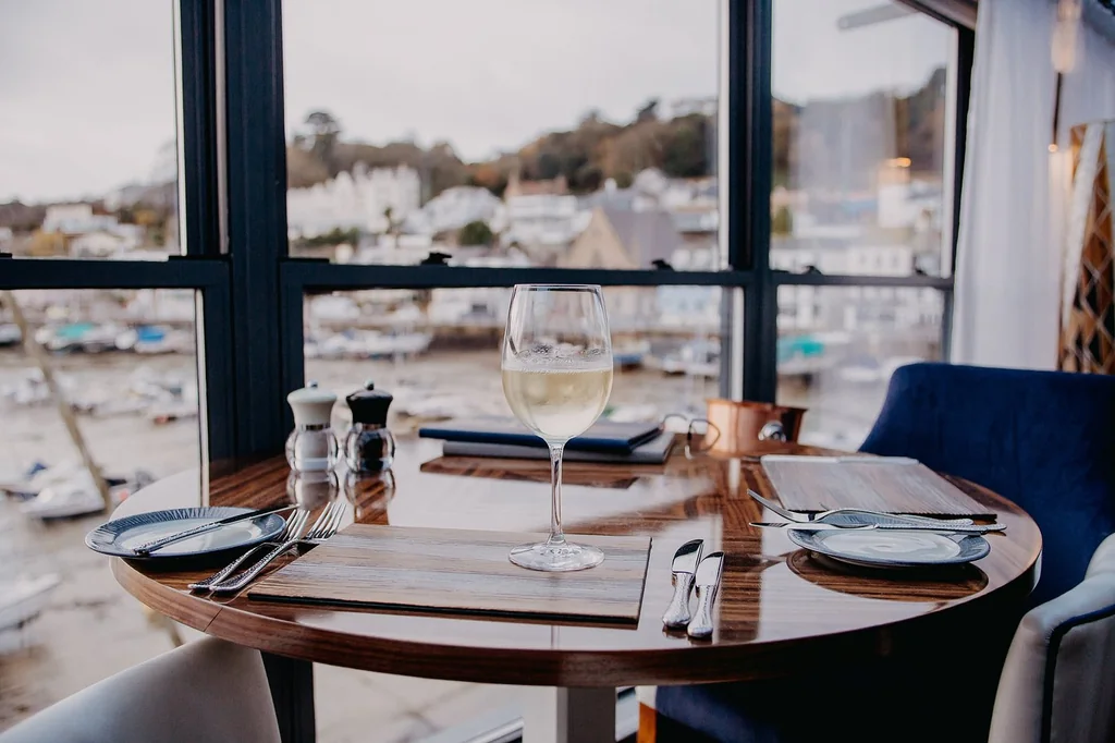 A serene dining scene is set on a wooden table with a white tablecloth, featuring a white wine glass, silverware, and a blue napkin. A salt and pepper shaker are on the left, and a pepper and salt shaker are on the right. The table is near a window with a view of a harbor and boats, creating a tranquil atmosphere.
