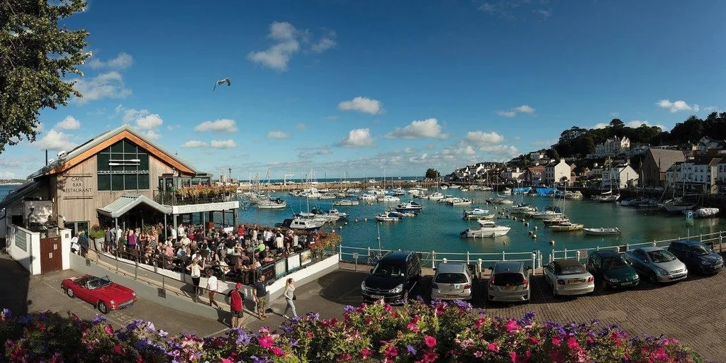 A bustling harbor scene features a large wooden building with a glass roof, surrounded by colorful flowers and a bustling pier filled with boats. A red car is parked nearby, and a bird soars above. The sky is a clear blue with scattered clouds, and the image is taken from a high vantage point, offering a panoramic view of the harbor and surrounding area.