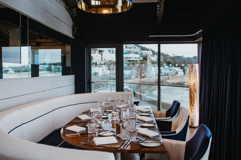 A modern restaurant interior features a curved wooden table with white napkins, silverware, and glasses, surrounded by white booths. The table is set for a formal dining experience, with a large window offering a view of a cityscape. The room is illuminated by a gold chandelier, creating a warm and inviting atmosphere.
