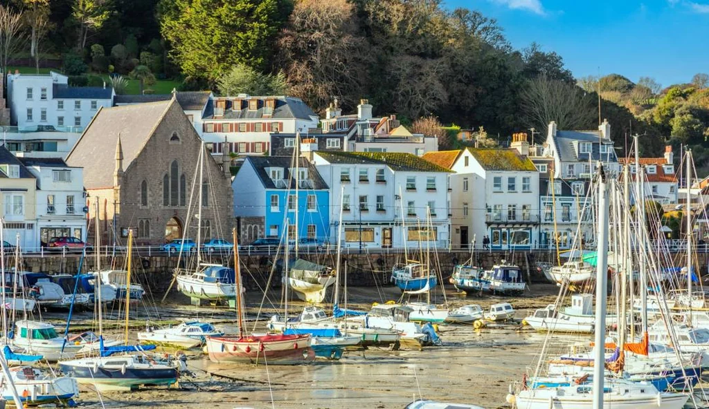The image features a serene harbor with 20 boats of various sizes and colors, docked in calm water. The boats are scattered across the harbor, with some closer to the viewer and others further away. The harbor is surrounded by a hill with white buildings and trees, creating a picturesque scene. The sky is clear blue with a few clouds, and the overall atmosphere is peaceful and idyllic.