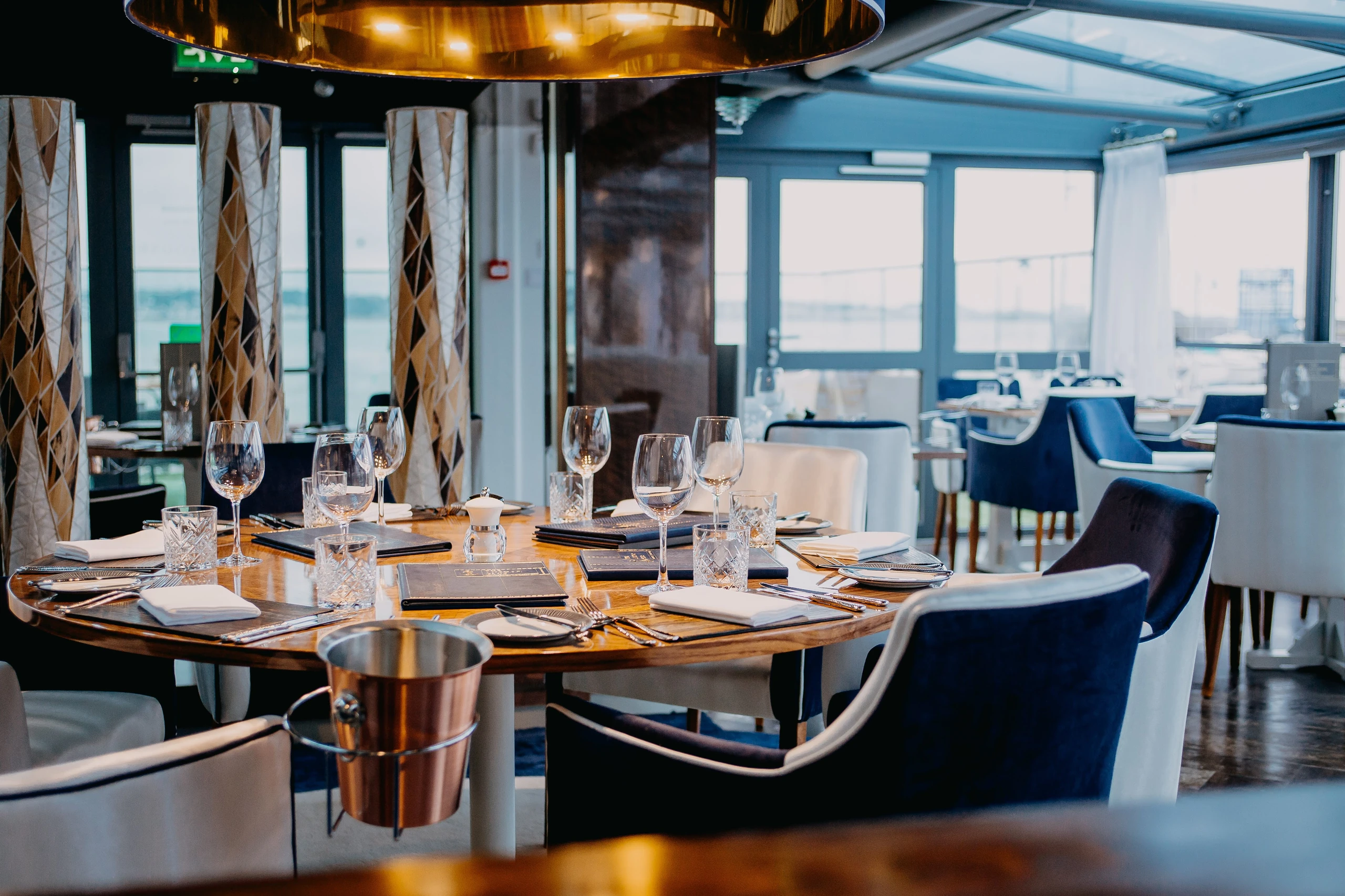 The image shows a modern restaurant with a wooden table set for a meal, surrounded by blue and white chairs. The table is adorned with silverware and wine glasses. The restaurant has large windows offering a view of the sea, and the floor is made of polished wood. The lighting is warm and inviting, creating a serene atmosphere. The landmark "sa_15007" is not identifiable in the image.