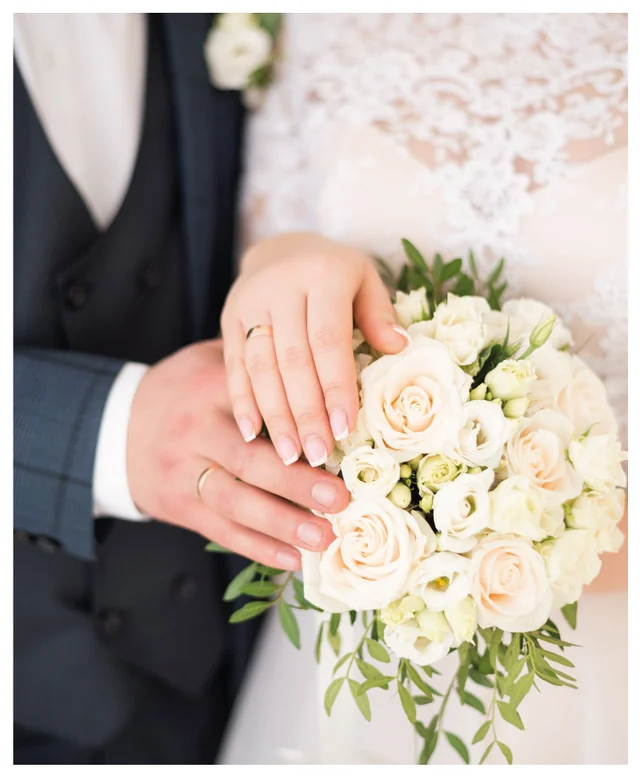 A couple in a black tuxedo and white wedding dress, holding hands and adorned with wedding rings, stands in a white background, creating a romantic and intimate atmosphere.
