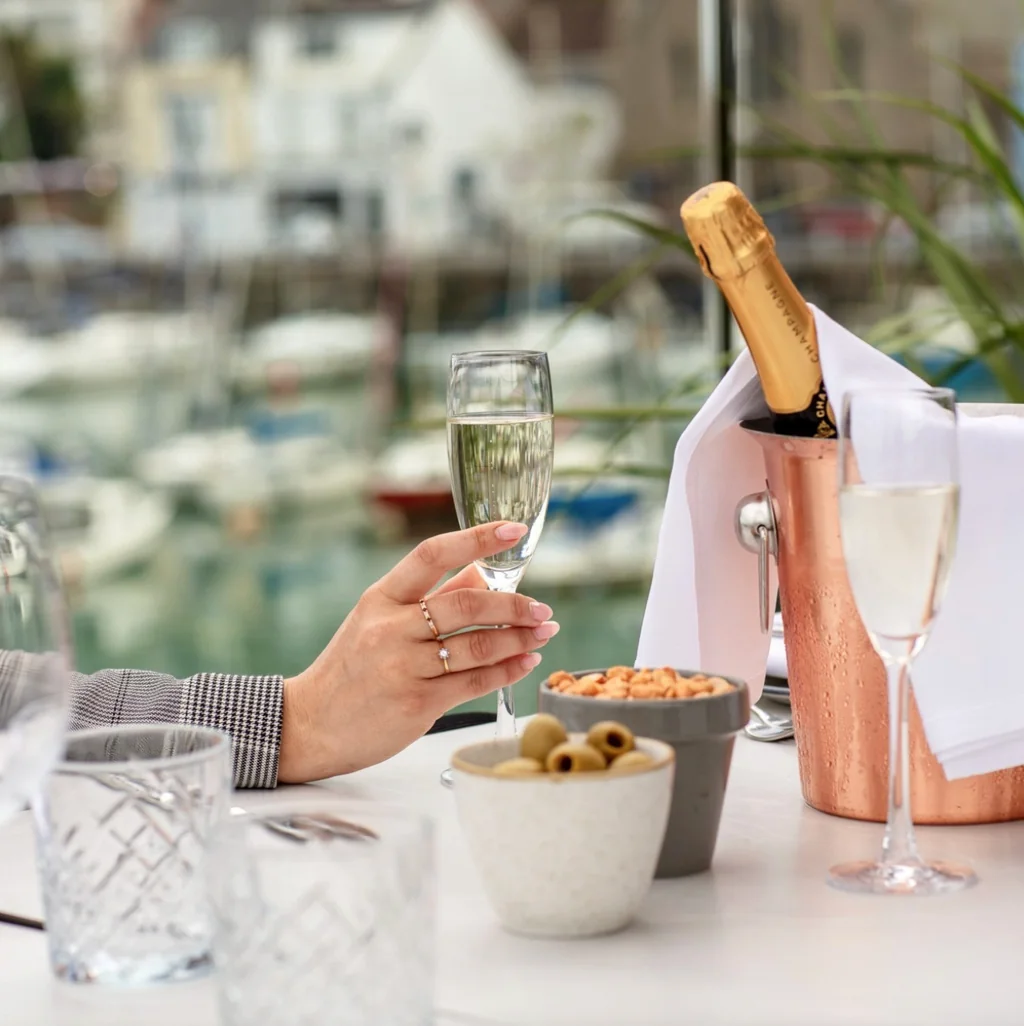 A serene dining scene features a table with a white tablecloth, a wine bottle with a golden cork, a bowl of olives, and a salt shaker. The background shows a marina with boats and a window overlooking the water. The image is taken from a low angle, emphasizing the table and objects. The scene suggests a romantic evening at a marina resort.