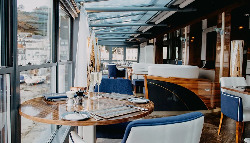 The image shows a modern restaurant with a wooden table set for a meal, surrounded by blue chairs and a white tablecloth. The restaurant has a glass ceiling and a view of a cityscape. The layout is well-planned, with tables and chairs arranged for optimal seating and a menu displayed for guests. The image is taken from the perspective of the restaurant's entrance, inviting viewers to imagine themselves dining there.