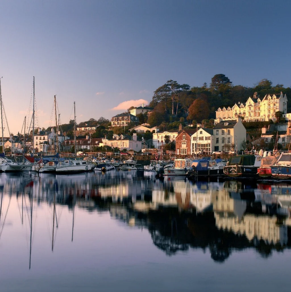 The image features a serene harbor scene with a clear blue sky, calm water, and various boats of different sizes and colors. The boats are docked in the harbor, reflecting in the water. The harbor is surrounded by a hill with colorful houses, and the sky is a light blue with scattered clouds. The scene is peaceful and tranquil, with no discernible text or human activity.