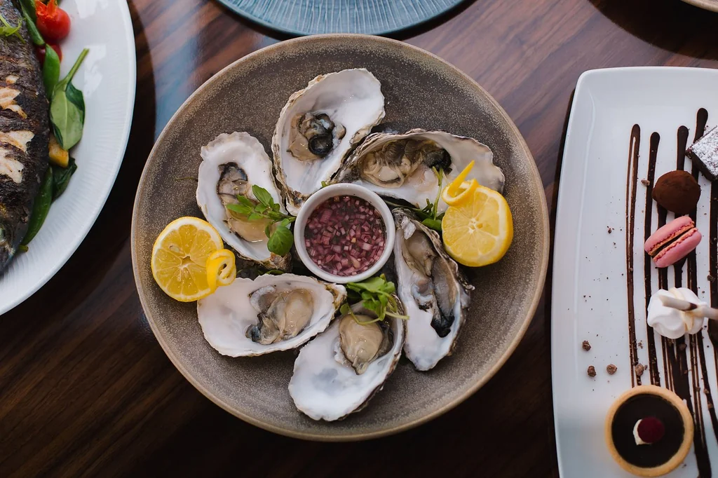 A dark brown plate holds six oysters, garnished with lemon slices and red sauce. A fork and knife are nearby, and a pink salmon dish is in the background. The scene is set on a wooden table, creating a luxurious dining atmosphere.