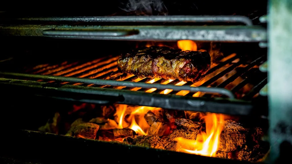 A close-up image shows a grill with a steak on a metal rack, burning with flames and charred meat. The grill is black, contrasting with the grill's metal structure. The steak is the main focus, with the grill's metal structure providing a sturdy base. The image captures a moment of cooking, showcasing the grill's functionality and the steak's deliciousness.