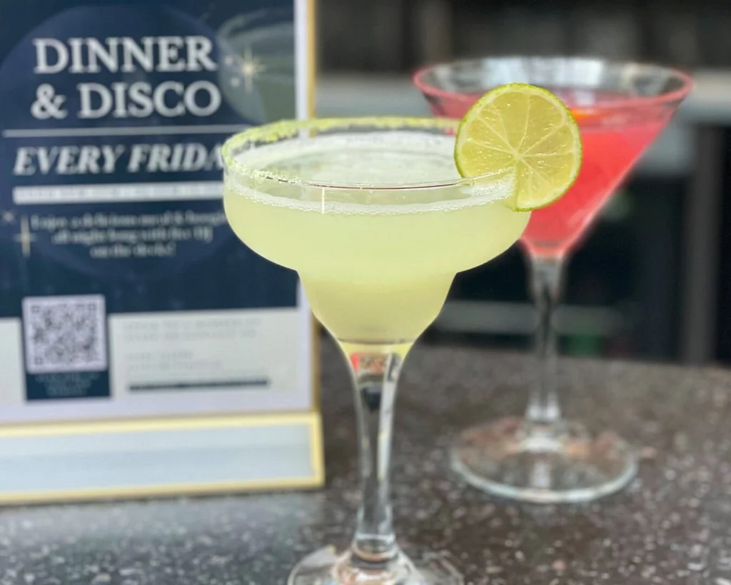Two cocktail glasses, one yellow and one pink, are on a black countertop. The yellow glass has a lime slice on top, while the pink glass has a white label. The background is blurred, focusing on the glasses and their contents.