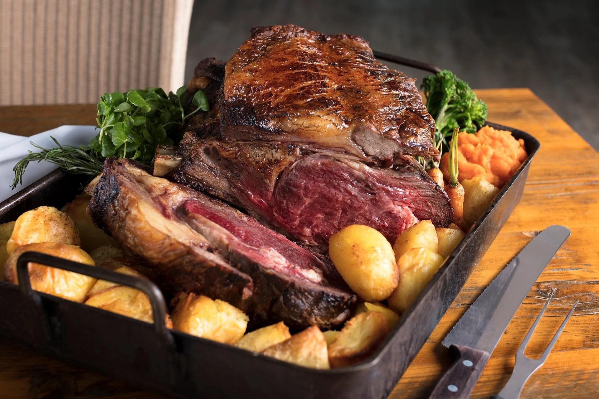 A large, well-cooked roast beef in a black tray is the centerpiece of a meal, accompanied by roasted potatoes, carrots, and a sprig of rosemary. The tray sits on a wooden table, with a fork and knife nearby, creating a warm and inviting atmosphere.