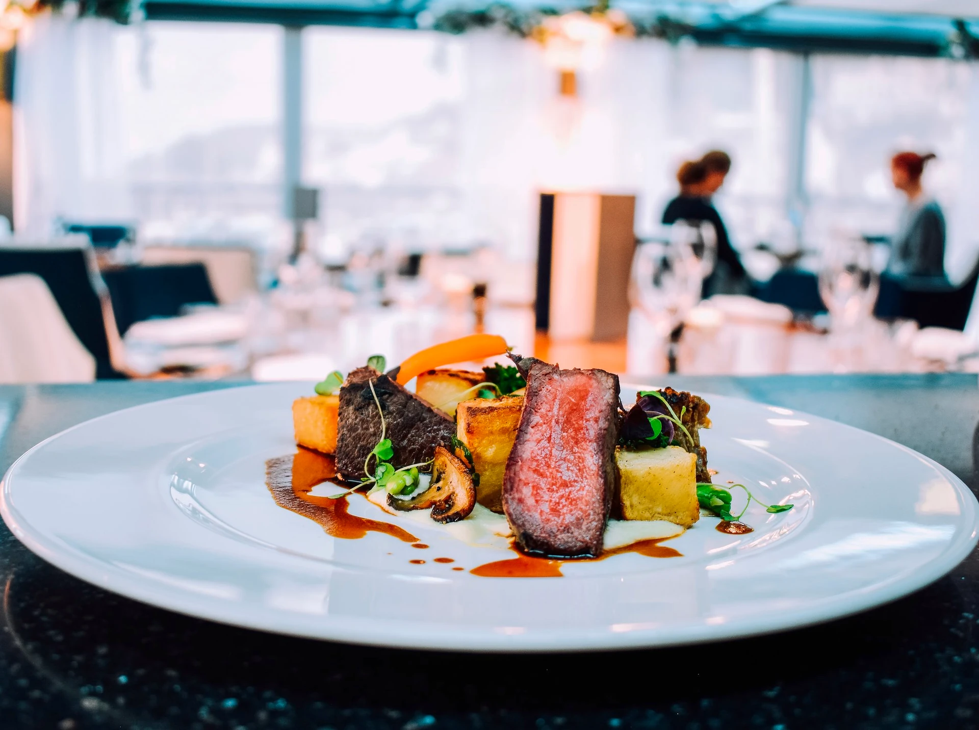 A white plate in a restaurant features a medium-rare steak with pink sauce, garnished with parsley and orange carrots, and served on a bed of potatoes. The plate is on a black tablecloth, with a large window in the background revealing a cityscape. The scene is visually appealing and suggests a high-end dining experience.