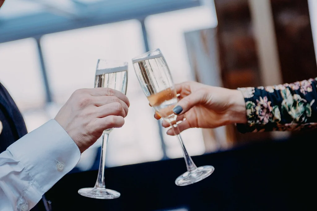 Two hands are holding champagne glasses, with one hand holding a small blue gemstone. The background is blurred, suggesting a cityscape or indoor setting. The focus is on the hands and glasses, with the gemstone adding a unique touch. The image conveys a sense of celebration and joy.
