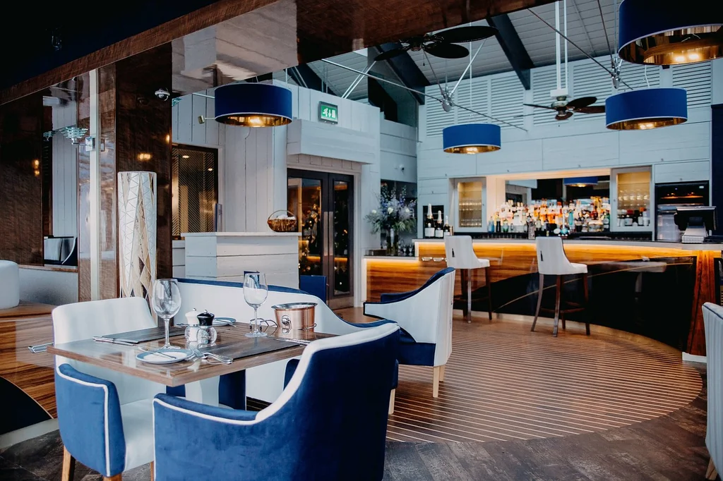 The image features a modern, minimalist restaurant with a wooden floor, white walls, and blue accents. The bar area has a counter with bar stools and a bar counter with a bar. The ceiling has a fan and pendant lights, and the space is well-lit. The layout is organized, with tables and chairs arranged for dining and socializing. The restaurant is ready for guests to enjoy a meal and conversation.