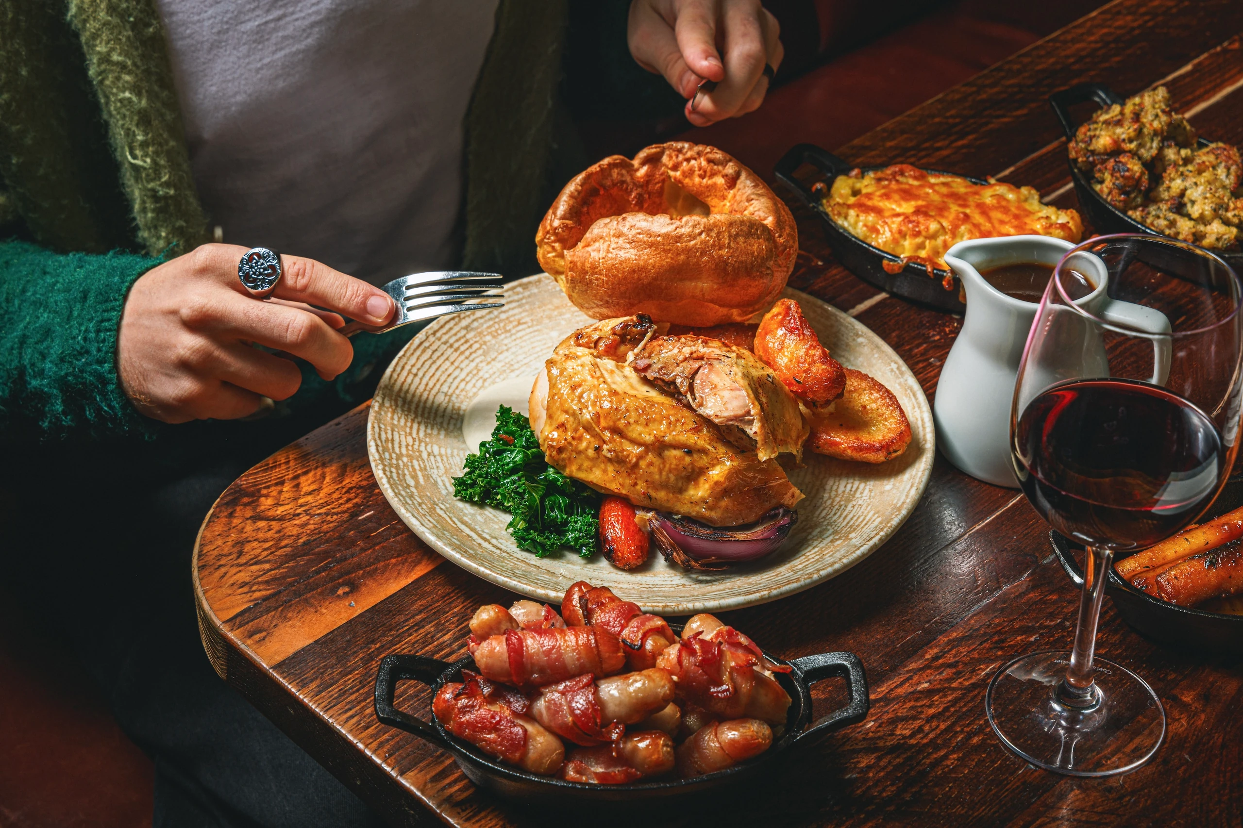 A person in a green sweater is seated at a wooden table, reaching for a large, golden-brown roast chicken sandwich with onions and red sauce. A plate of roasted potatoes and a pitcher of red wine are nearby. A white pitcher with a red liquid is also present. The scene is set against a dark background, emphasizing the main subject and its surroundings.