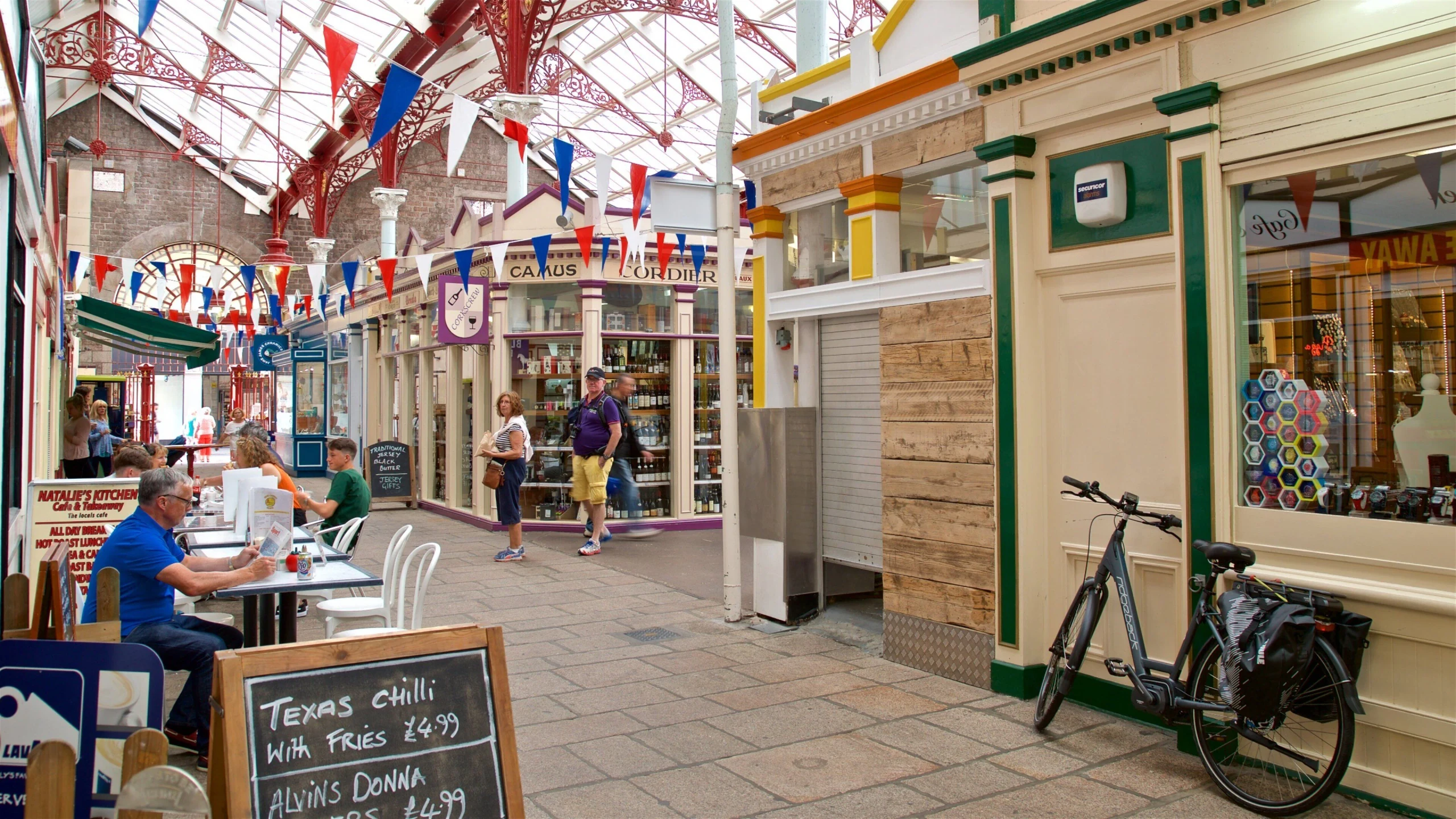 The image shows a lively shopping center with a glass ceiling, red and white flags, and colorful shops. People are walking around, some seated at tables, and a black bicycle is parked nearby. A chalkboard menu is displayed, and a chalkboard sign is visible. The scene is bustling and vibrant, with a mix of people and objects.