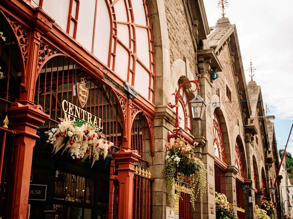 The image features a charming, red-bricked building with a large arched entrance and a coat of arms above. The building is adorned with hanging flowers and a clock tower, and is located on a street lined with other buildings. The sky is overcast, casting a soft light on the scene. The image captures the essence of a bustling city, with the building as the focal point.