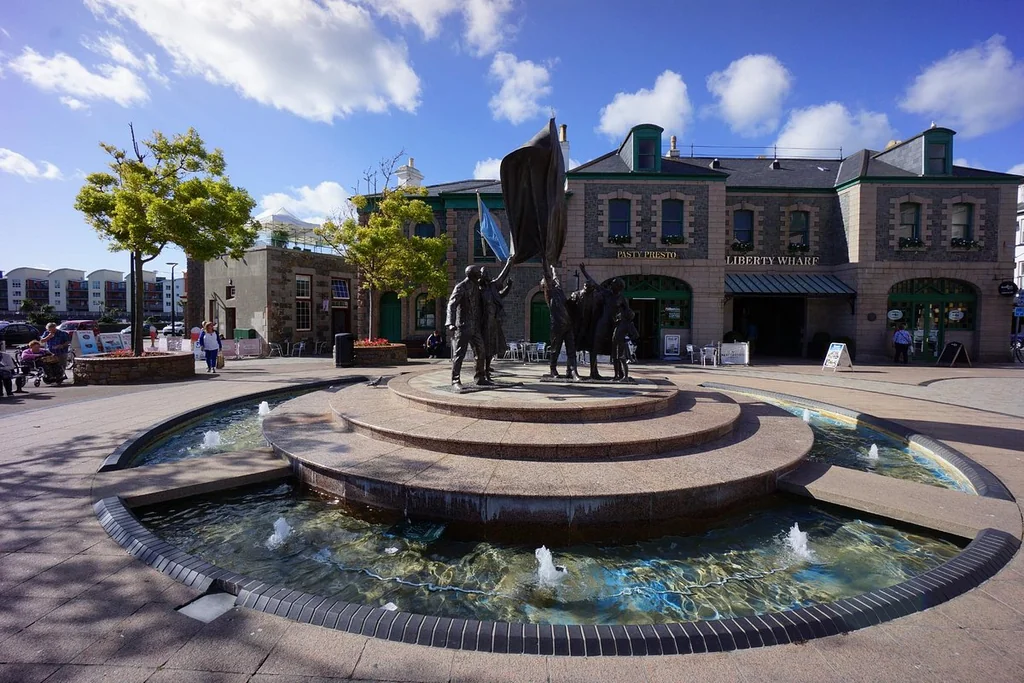 A large, black metal sculpture of a man holding a flag stands in a circular stone fountain, surrounded by people. The fountain has four water jets, and the scene is set against a backdrop of buildings and trees. The image is taken from a low angle, emphasizing the sculpture's grandeur.