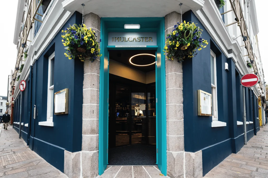 The image shows the entrance of the "Hermel & Hutterley" store in London, UK, with a blue facade and a green door. Two hanging flower baskets with yellow and blue flowers are above the door. The street is lined with buildings and a red "No Parking" sign is visible. The scene is lively and inviting, with the store's name and location prominently displayed.