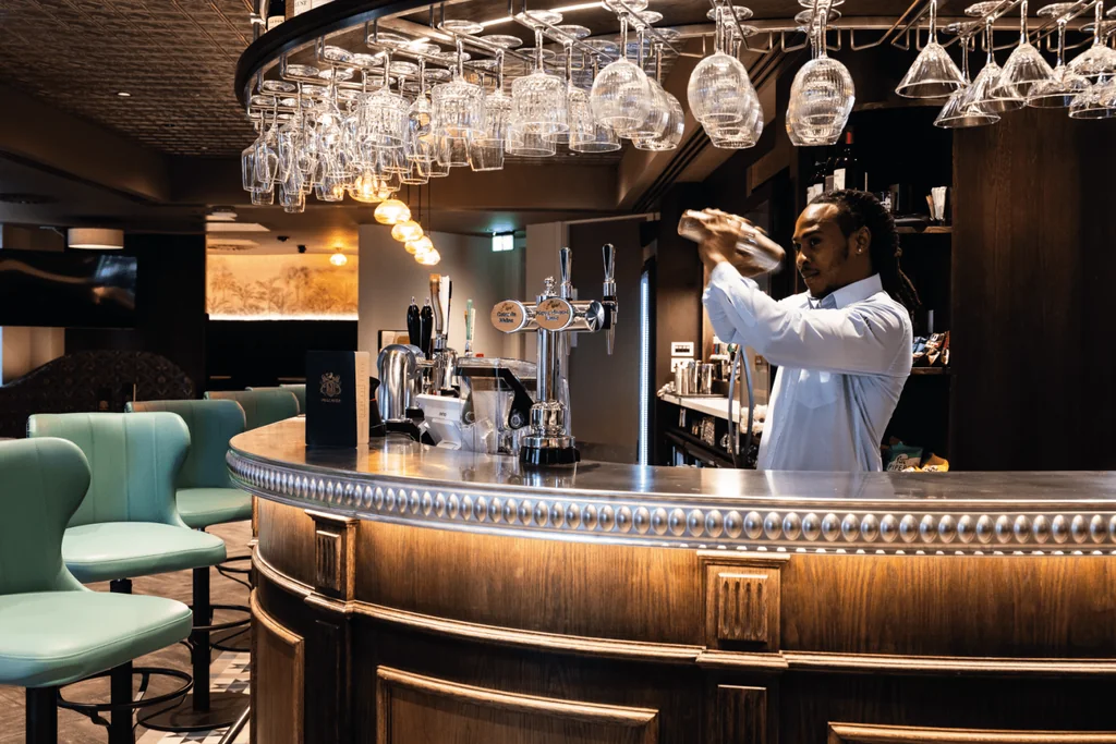 In a modern bar, a bartender in a white shirt pours a clear liquid into a glass at the bar's center. The bar features a polished wooden counter, a glass display, and a blue and green bar stool. The background includes a brick wall, a hanging light fixture, and a black and white rug. The scene is illuminated by a warm, inviting atmosphere.