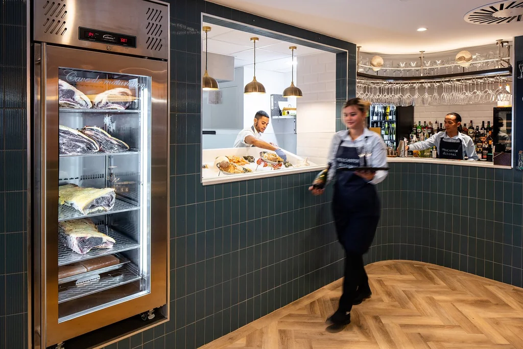 The image depicts a modern, well-lit kitchen with a large refrigerator filled with fresh seafood, a counter with a man preparing food, and a man in a striped shirt and apron working behind the counter. The walls are covered in blue tiles, and the ceiling features hanging lights. The scene is bustling and inviting, with a sense of order and efficiency.