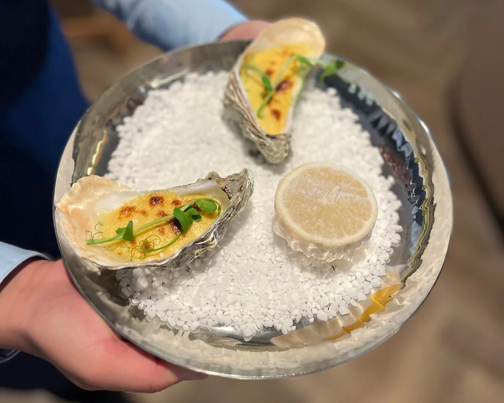 A silver tray with three oysters on white rice is held by a hand, with a lemon slice on top. The oysters are golden brown, with green herbs and a crispy crust. The background is blurred, focusing on the tray and oysters.