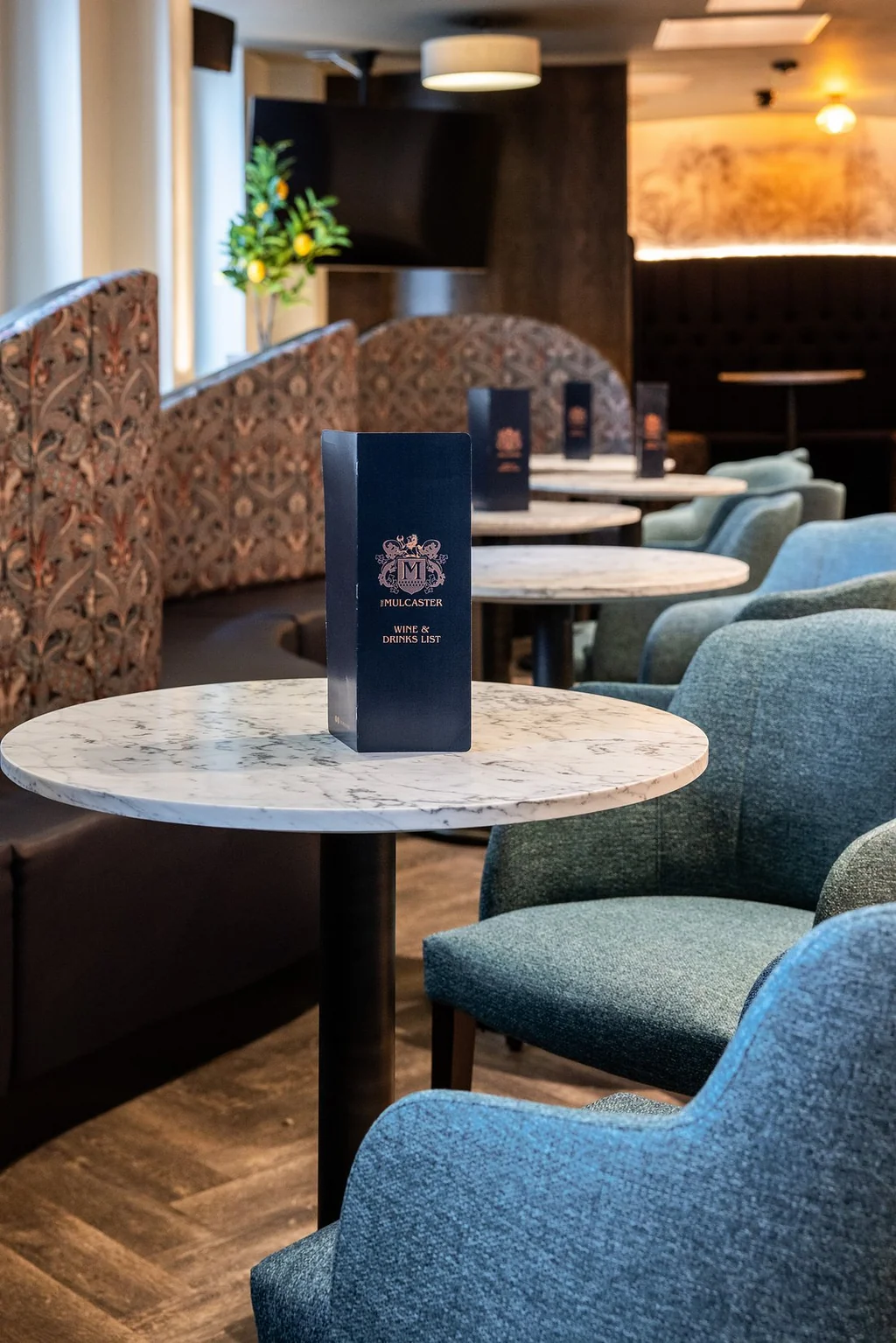 The image features a modern coffee shop with a marble table, a black "Café du Sud" menu, and blue armchairs. The background has a floral patterned wall and a wooden ceiling. The perspective is from the front, looking towards the back, creating a sense of depth and inviting customers to sit and enjoy their coffee.