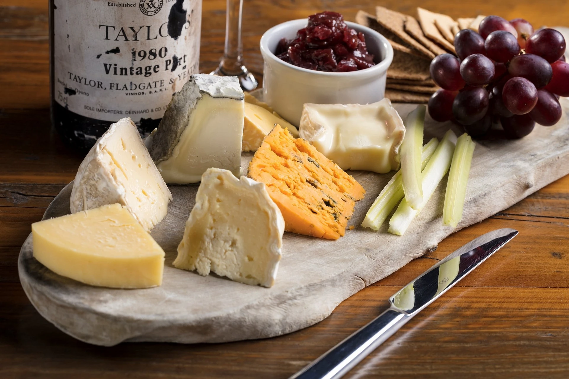 A rustic wooden table features a wine bottle with "Tirone" label, a white wine glass, a cheese board with various cheeses, grapes, and cranberries, a knife, and a bowl of red jam. The arrangement creates a harmonious and inviting atmosphere.