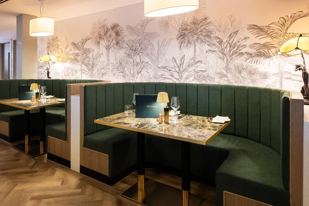 The image shows a modern, minimalist restaurant with a neutral color scheme, featuring a green and white striped wallpaper, a wooden floor, and a wooden bench with green seats. The tables are set with white napkins, glasses, and silverware, and are accompanied by white lamps. The arrangement of furniture and decor creates a harmonious and inviting atmosphere.