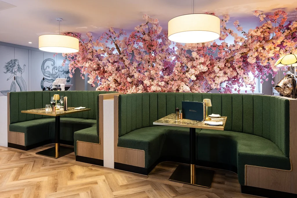 The image features a modern restaurant with a vibrant floral wall, green booths, and a wooden floor. The booths are adorned with green velvet seats and white tablecloths, while the floor is made of light wood. The walls are decorated with pink and white flowers, and the ceiling has white light fixtures. The restaurant is well-lit and offers a cozy atmosphere.