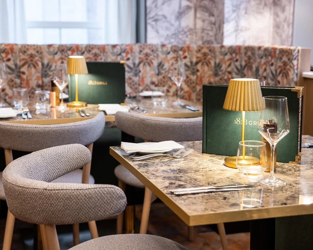 The image features a modern restaurant with a marble table, four gray chairs, and a green menu. A gold lamp with a yellow shade is on the left side, casting a warm glow. The background has floral wallpaper and a window with white curtains. The arrangement of objects creates a harmonious and inviting atmosphere.