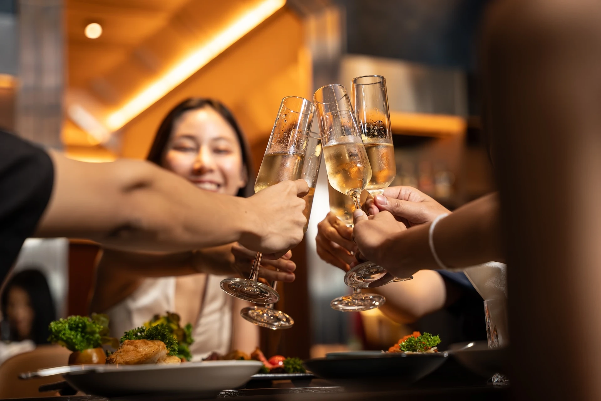 A lively gathering of people, dressed in white and black, are joyfully raising their glasses in celebration. The glasses are filled with a golden liquid, possibly champagne, and are held by their hands. The scene takes place on a table with a plate of food and a salad, surrounded by a warm, inviting atmosphere. The background is blurred, emphasizing the joyous moment.
