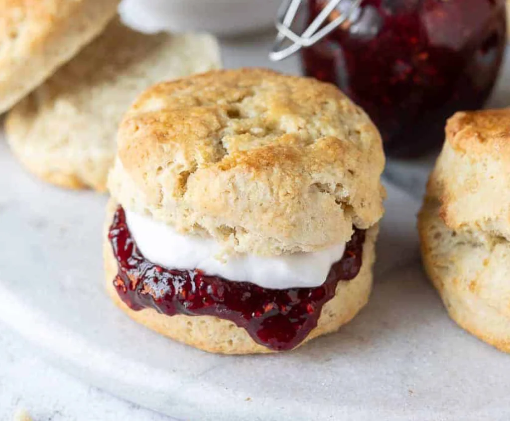 A white plate holds three scones of scones, each with a cream-filled sandwich. The scones are golden brown, with a creamy filling. A jar of jam is in the background, and a silver wire rack is nearby. The scene is simple and inviting, with the scones as the main focus.