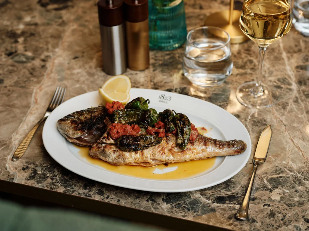 A white plate with a golden rim holds a grilled fish with a lemon slice, garnished with red and green vegetables, on a marble table. A silver fork and knife are nearby, and a wine glass and a champagne flute are on the table. A gold salt shaker and a gold napkin holder are in the background. The scene is elegant and sophisticated.