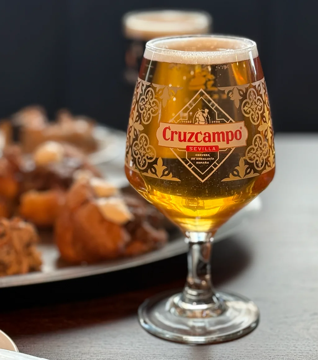 A glass of Cavaçort beer, with a golden amber color and a red and white label, is the main subject in the image. It is placed on a dark table, with a plate of pastries in the background. The background is blurred, focusing on the beer and pastries. The image is a still life, with no movement or action.