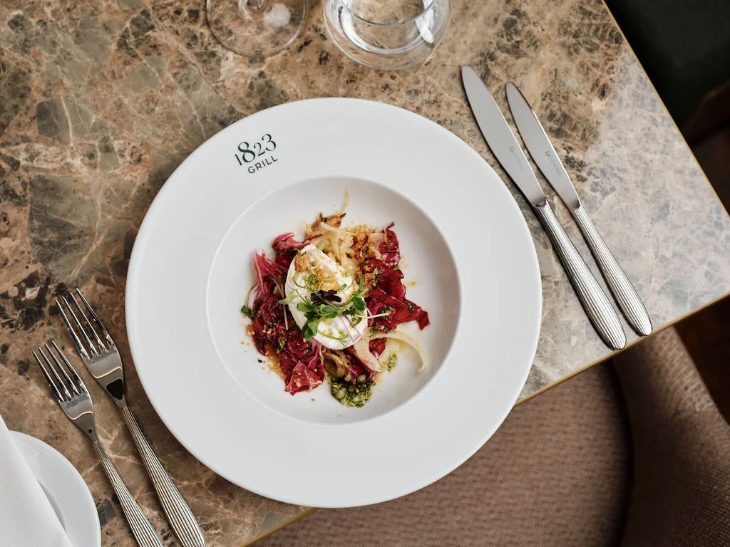 A white plate with a vibrant red and green salad, garnished with greens and a fried egg, is the centerpiece of a dining table. Two silver forks and knives are positioned to the left and right of the plate, with a wine glass to the right. The table is set against a dark marble countertop, creating a sophisticated and elegant atmosphere.