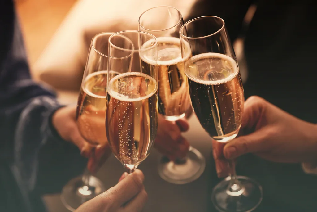 Six champagne glasses filled with bubbly champagne are being raised in a joyful toast, with each glass slightly tilted and filled with bubbles. The background is blurred, emphasizing the toast and the glasses. The image captures a moment of celebration and camaraderie.