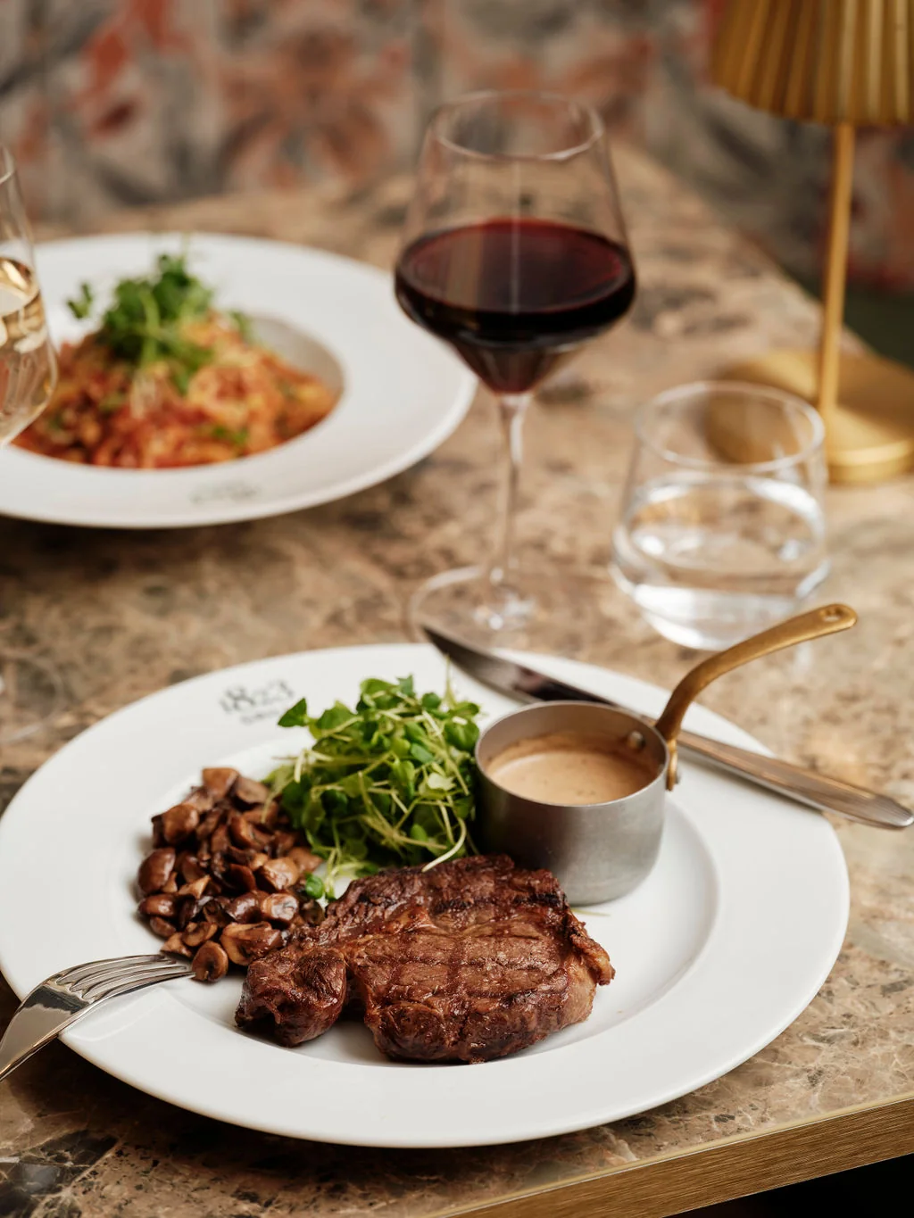 A luxurious dining scene features a white plate with a steak, salad, and sauce, accompanied by two glasses of red wine and a silver fork and knife. A gold-colored lamp adds elegance, and the table is covered with a floral pattern. The image portrays a sophisticated dining experience.