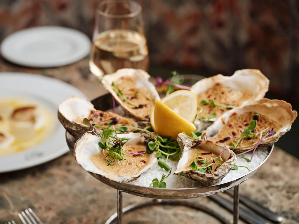 A silver tray on a wooden table holds six oysters, garnished with fresh herbs and a lemon slice. A glass of white wine and a plate of seafood are in the background, creating a sophisticated dining atmosphere.