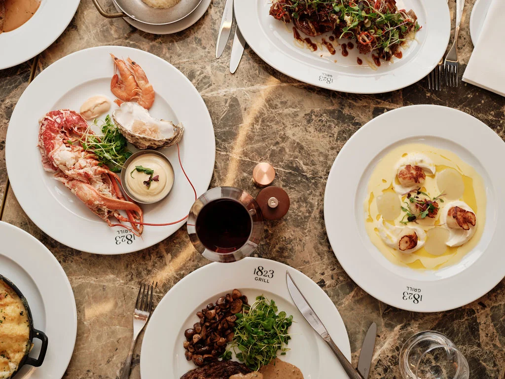 The image features a dining table with six plates of food, each with a unique dish. The top left plate has a lobster, the top right plate has a seafood dish, the bottom left plate has a seafood dish, the bottom right plate has a seafood dish, and the bottom left plate has a seafood dish. The table is set on a marble tablecloth, with a wine glass and a fork nearby. The arrangement creates a visually appealing and appetizing scene.