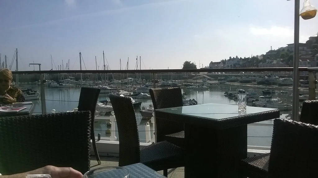 A serene outdoor cafe on a balcony overlooks a bustling harbor filled with boats. The cafe features a glass table with a clear glass top, a black metal railing, and black wicker chairs. A person is seated at the table, and the background shows a picturesque harbor with boats and buildings. The scene is bathed in natural light, creating a tranquil atmosphere.