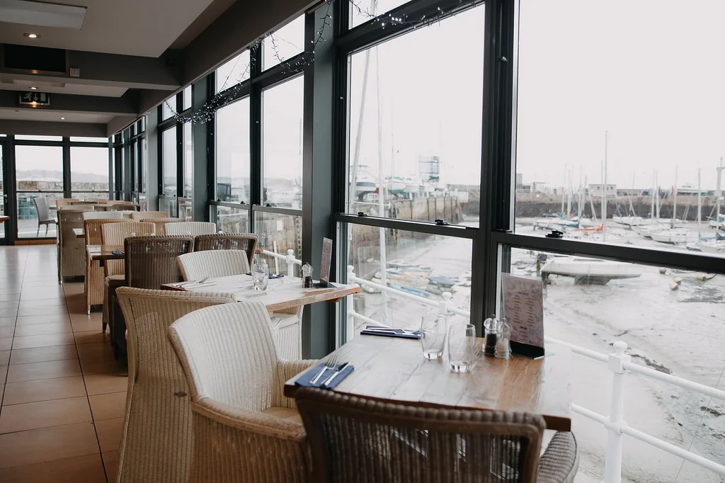 The image shows a modern restaurant with large windows offering a view of a harbor with boats. The restaurant has a warm, inviting atmosphere with wooden tables and chairs, white curtains, and blue napkins. The perspective is from the front of the restaurant, looking towards the harbor, creating a sense of depth and inviting customers to enjoy the view.