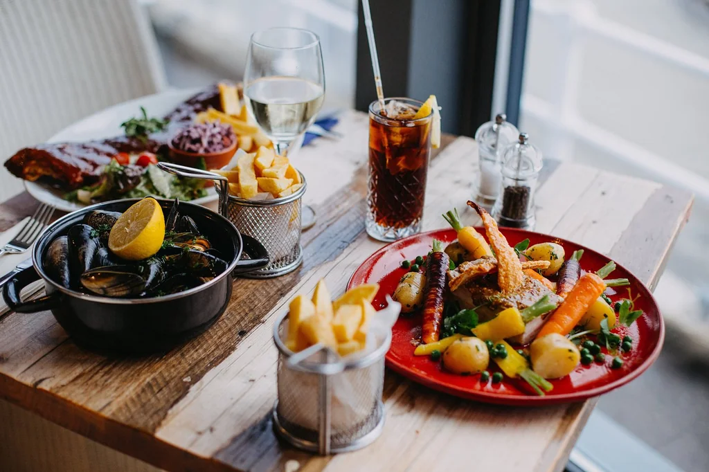 A wooden table is set for a meal, with a black pot filled with mussels and a white plate of potatoes and vegetables. A glass of iced tea with a straw is on the left, and a glass of water with a straw is on the right. The background shows a window with a view of a building, creating a cozy atmosphere.
