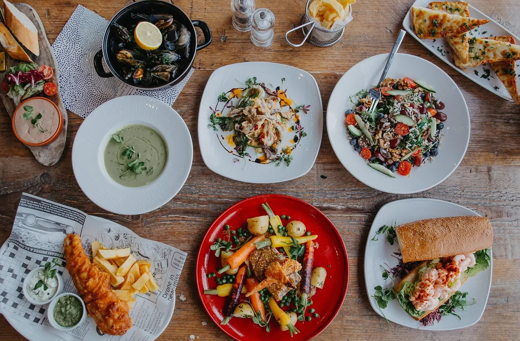 The image features a rustic wooden table with six plates of diverse food, arranged in a circular pattern. The plates contain a variety of dishes, including pasta, seafood, salad, soup, and fried chicken. The table is adorned with condiments, a salt shaker, and a pepper grinder. The arrangement creates a visually appealing and appetizing scene.