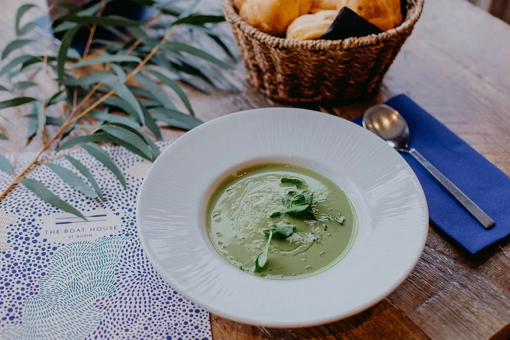 A serene dining scene features a white plate with a green soup, garnished with parsley, on a wooden table. A blue and white placemat holds bread and a basket of fresh green leaves. A spoon is nearby, and the blurred background suggests a cozy, rustic setting. The image captures a moment of tranquility and anticipation for a meal.