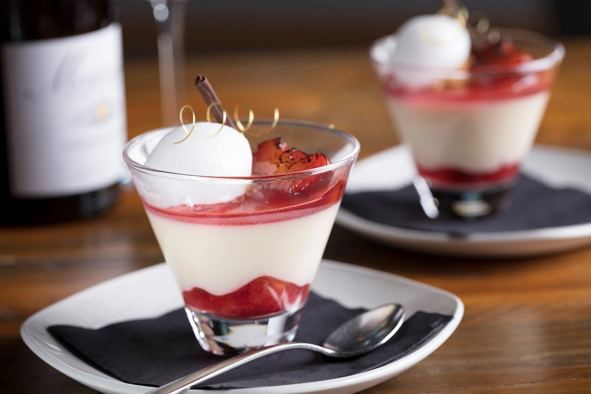 Two glass dishes with white and red desserts, topped with strawberries and a small gold decoration, are on a wooden table. A spoon is on the left dish, and a wine bottle is in the background. The scene is visually appealing and suggests a delightful dessert experience.