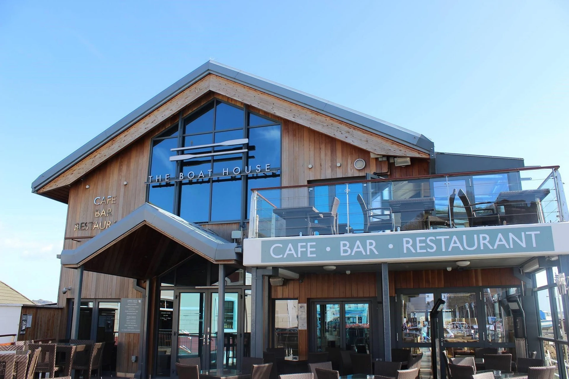 The image features the modern, wooden building of the "Carn Bar & Grill" restaurant, with a sloping roof and large glass windows reflecting the blue sky. The name is displayed in white letters above the entrance, and the restaurant is located on a pier with outdoor seating and a deck. The image is taken from a low angle, emphasizing the building's grandeur and inviting atmosphere.