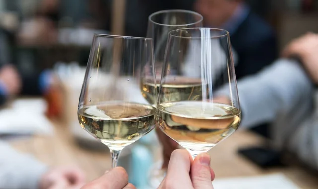 Two hands raise wine glasses filled with white wine, creating a toast-like gesture. The blurred background suggests a social gathering, possibly a restaurant or bar. The focus is on the wine glasses and the hands, with no text or other objects present.