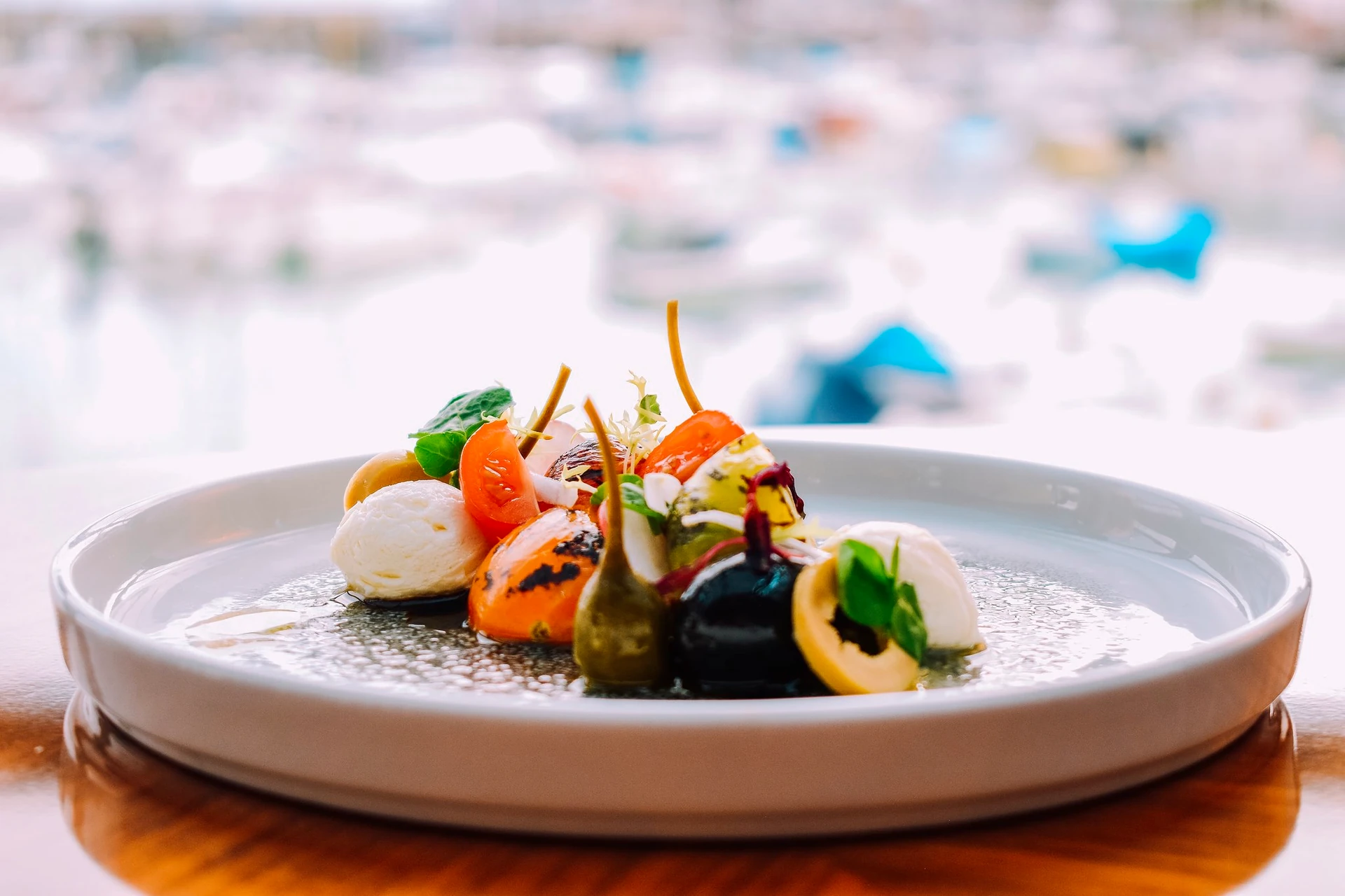A white plate with a colorful, appetizing seafood dish is on a wooden table, surrounded by a blurred background of boats and buildings. The dish features a variety of colorful vegetables, including orange, red, and green, and is garnished with basil leaves. The plate is set on a wooden surface, creating a rustic and elegant atmosphere.